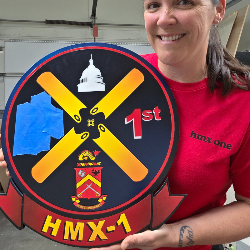 Person holding a large round wood emblem with '1st 1st HMX-1' text and symbols.